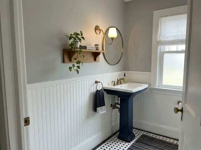 Small powder room with a navy vanity, round brass mirror and white wainscoting